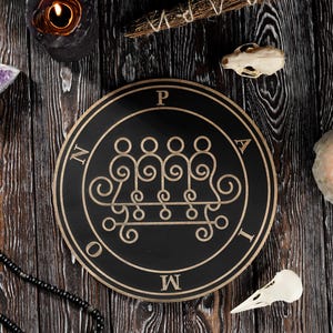 Pode incluir: Um emblema circular preto e dourado com figuras estilizadas e texto, rodeado por velas, cristais e um crânio de pássaro, sobre um fundo de madeira escura. O emblema tem as letras P, A, T, M, N e I.