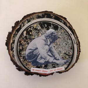 May include: A round wooden slice with a black and white city map design. A figure of a person is kneeling in the center of the map. The text "We cannot choose not to choose." is printed on a white label below the figure.