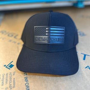 May include: A black baseball cap with a black and white American flag patch and the text "ONE NATION UNDER GOD".