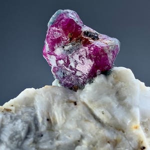Może przedstawiać: Surowy okaz kryształu w kolorze głębokiego magenty, osadzony w teksturowanej, złamanej bieli matrycy mineralnej. Kryształ posiada nieregularne, fasetowane powierzchnie z widocznymi inkluzjami oraz błyszczący, półprzezroczysty wygląd na matowym tle.