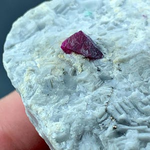 Può includere: Primo piano di una roccia grigio chiaro con un cristallo rosso scuro incorporato. Il cristallo è angolare e riflette la luce. La roccia ha una superficie strutturata con piccole macchie di altri minerali. Il cristallo è rosso intenso.