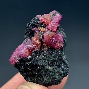 May include: A close-up of a raw ruby specimen. The ruby crystals are a vibrant pinkish-red colour, contrasting with the dark, rough matrix. The mineral formation has a natural, unpolished appearance, showcasing the raw beauty of the gemstone.