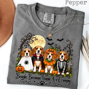 May include: A pepper-coloured t-shirt featuring a graphic of four beagles in Halloween costumes, including a ghost, vampire, pumpkin, and skeleton. The design includes a full moon, bats, and the text "Beagle, Because People Are Creepy" in black.