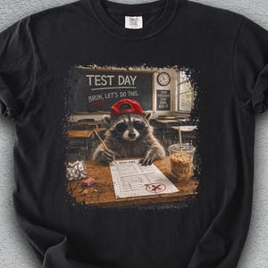 May include: Black t-shirt with a raccoon wearing a red cap, taking a test in a classroom. The text "TEST DAY BRUH, LET'S DO THIS" is above the raccoon. A cup of coffee and a crumpled paper are on the desk.