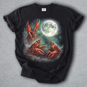 K&ouml;nnte beinhalten: Schwarzes T-Shirt mit einer surrealen Illustration von drei roten Krebsen vor einem Vollmond. Die Krebse sind so positioniert, als w&uuml;rden sie tanzen oder interagieren, mit einem Sternenhimmel-Hintergrund.