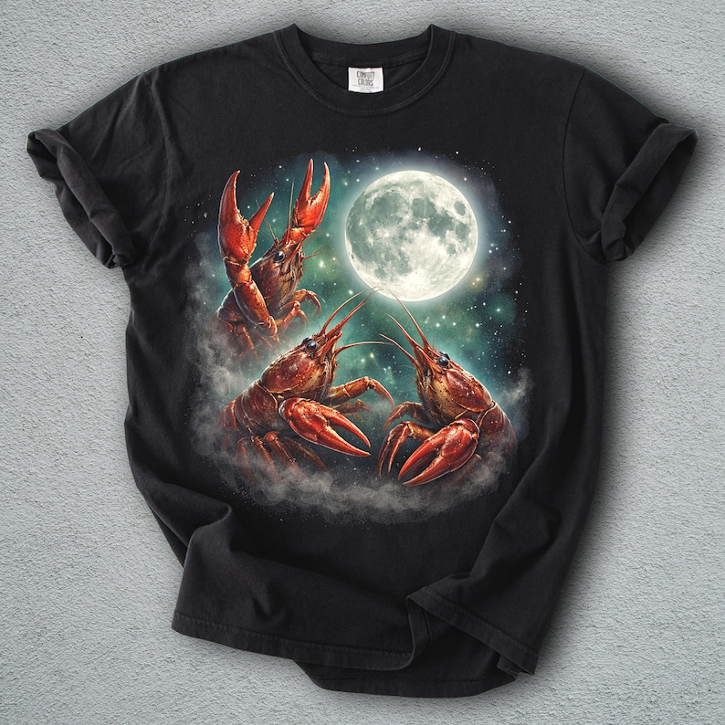 K&ouml;nnte beinhalten: Schwarzes T-Shirt mit einer surrealen Illustration von drei roten Krebsen vor einem Vollmond. Die Krebse sind so positioniert, als w&uuml;rden sie tanzen oder interagieren, mit einem Sternenhimmel-Hintergrund.