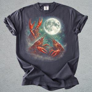 K&ouml;nnte beinhalten: Dunkelgraues T-Shirt mit einer Illustration von drei roten Krebsen vor einem Vollmond und einem Sternenhimmel. Das T-Shirt hat einen Rundhalsausschnitt und kurze &Auml;rmel. Das Design ist detailliert.