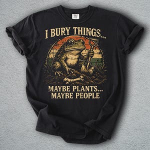 Pode incluir: T-shirt preto com um desenho engraçado de um sapo segurando uma pá. O texto diz: "I BURY THINGS... MAYBE PLANTS... MAYBE PEOPLE."