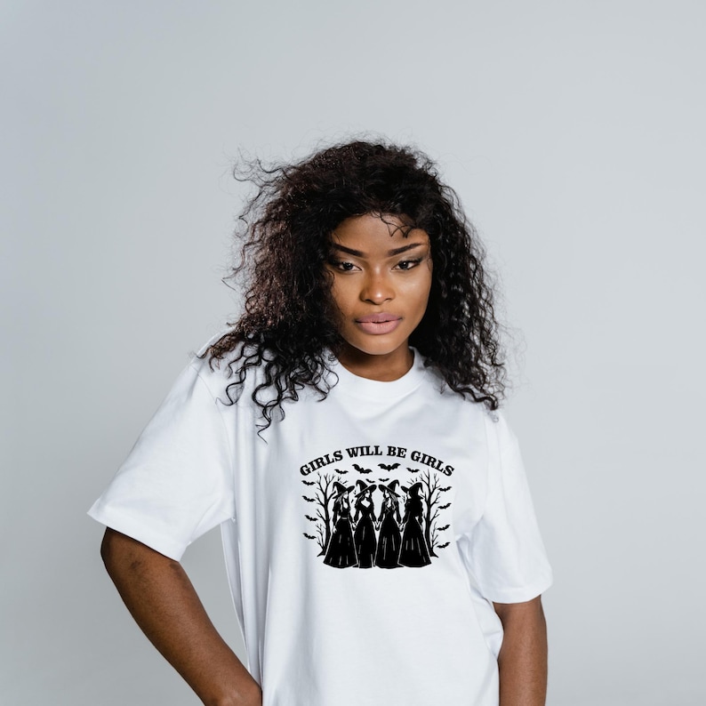 Puede incluir: Camiseta blanca con el texto negro "GIRLS WILL BE GIRLS". El dise&ntilde;o presenta siluetas de cuatro figuras con vestidos largos, murci&eacute;lagos y &aacute;rboles. La camiseta la lleva una persona con pelo rizado oscuro.