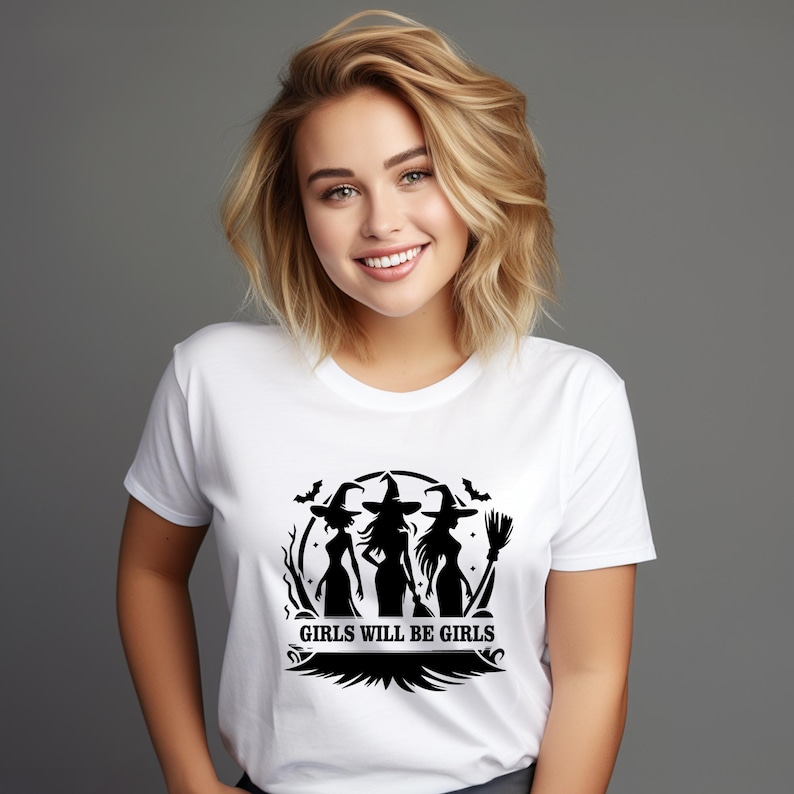 Puede incluir: Camiseta blanca con un gr&aacute;fico negro de tres siluetas de brujas, murci&eacute;lagos y una escoba. El texto "GIRLS WILL BE GIRLS" est&aacute; debajo. La camiseta la lleva una persona con pelo rubio.
