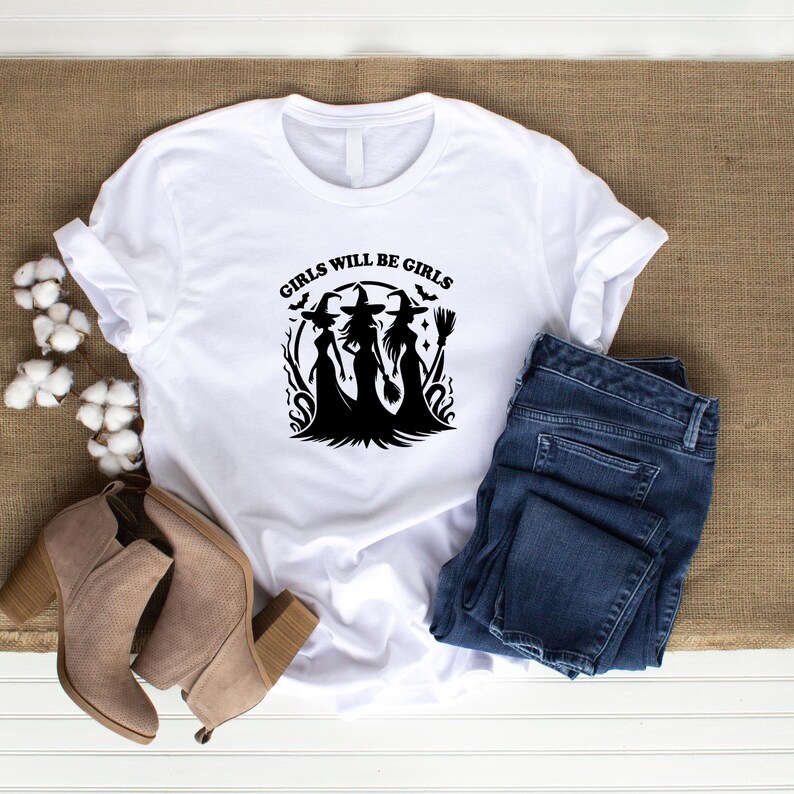 Puede incluir: Camiseta blanca con el texto "GIRLS WILL BE GIRLS" sobre un gr&aacute;fico negro de tres brujas. La imagen incluye vaqueros azules y botines color canela, sugiriendo un atuendo informal con tem&aacute;tica oto&ntilde;al.