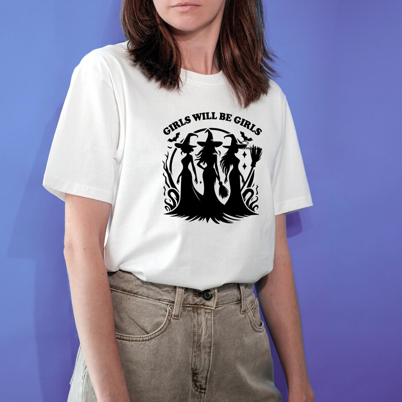 Puede incluir: Camiseta blanca con un gr&aacute;fico negro de tres siluetas de brujas y el texto "GIRLS WILL BE GIRLS". Las brujas llevan sombreros puntiagudos y sostienen una escoba. El dise&ntilde;o est&aacute; rodeado de remolinos decorativos y murci&eacute;lagos.