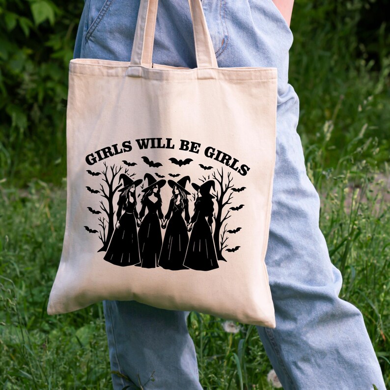 Puede incluir: Bolso tote beige con el texto negro "GIRLS WILL BE GIRLS". El dise&ntilde;o presenta siluetas de cuatro brujas, murci&eacute;lagos y &aacute;rboles desnudos. El bolso lo lleva una persona con vaqueros.