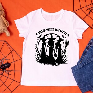Puede incluir: Camiseta blanca con un dise&ntilde;o gr&aacute;fico negro de tres brujas con sombreros puntiagudos y una escoba. El texto "GIRLS WILL BE GIRLS" est&aacute; arriba. Ropa con tem&aacute;tica de Halloween.
