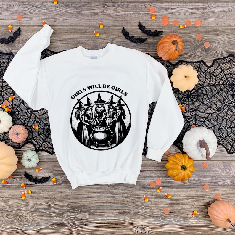 Puede incluir: Sudadera blanca con la frase "GIRLS WILL BE GIRLS" y un gr&aacute;fico en blanco y negro de tres brujas reunidas alrededor de un caldero. La sudadera est&aacute; rodeada de decoraciones de Halloween, incluyendo calabazas y murci&eacute;lagos.