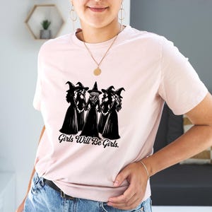 Puede incluir: Camiseta rosa claro con un dise&ntilde;o en blanco y negro de cuatro brujas tomadas de la mano. El texto "Girls Will Be Girls" est&aacute; impreso debajo. La camiseta est&aacute; hecha de un tejido suave.