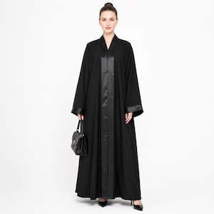 Puede incluir: Una abaya negra de cuerpo entero con mangas largas que presentan puños de satén y un panel vertical de satén a juego en la parte central delantera. La prenda se combina con un pequeño bolso de mano negro estructurado con asa superior.