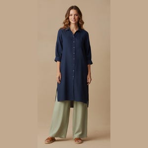 Puede incluir: Un vestido camisero largo azul marino con botones, mangas tres cuartos y un bolsillo en el pecho. El vestido se combina con pantalones de pierna ancha verde claro y sandalias marrones. El conjunto se presenta sobre un fondo neutro.