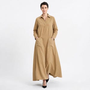 May include: A long-sleeved, floor-length tan dress featuring a collared V-neckline, a buttonless placket, and two large patch pockets on the front. The garment has a loose, flowing silhouette with a straight hemline and a solid, uniform color.