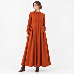 Puede incluir: Un vestido largo hasta el suelo de manga larga en un color naranja quemado sólido. La prenda presenta un cuello mao alto, una tapeta de botones frontal completa y un corpiño ajustado que se transforma en una falda plisada y voluminosa.