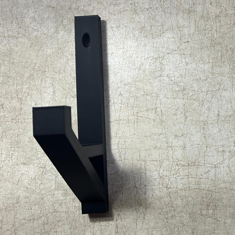 Puede incluir: Un gancho de pared negro. El gancho tiene un dise&ntilde;o rectangular con un soporte vertical y un brazo horizontal. Se perfora un agujero cerca de la parte superior para el montaje. El gancho est&aacute; hecho de un material negro mate.