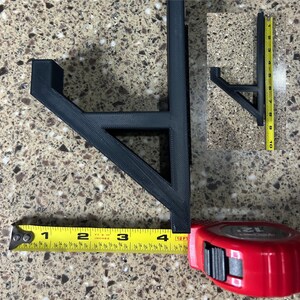 Puede incluir: Un soporte negro impreso en 3D, de aproximadamente 10 cm de largo, se muestra con una cinta m&eacute;trica. El soporte tiene un dise&ntilde;o en &aacute;ngulo recto con un soporte horizontal y un soporte vertical. Tambi&eacute;n se ve una versi&oacute;n m&aacute;s peque&ntilde;a del soporte.