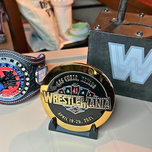 May include: A gold-colored coin with a black background featuring the text "Wrestlemania" and "Las Vegas, Nevada" with playing cards and the number 41. The coin is on a black stand.