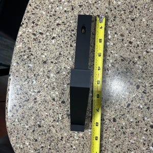 Puede incluir: Un soporte met&aacute;lico negro de forma rectangular, de aproximadamente 20 cm de largo, con un agujero en la parte superior. Una cinta m&eacute;trica amarilla se coloca junto al soporte.