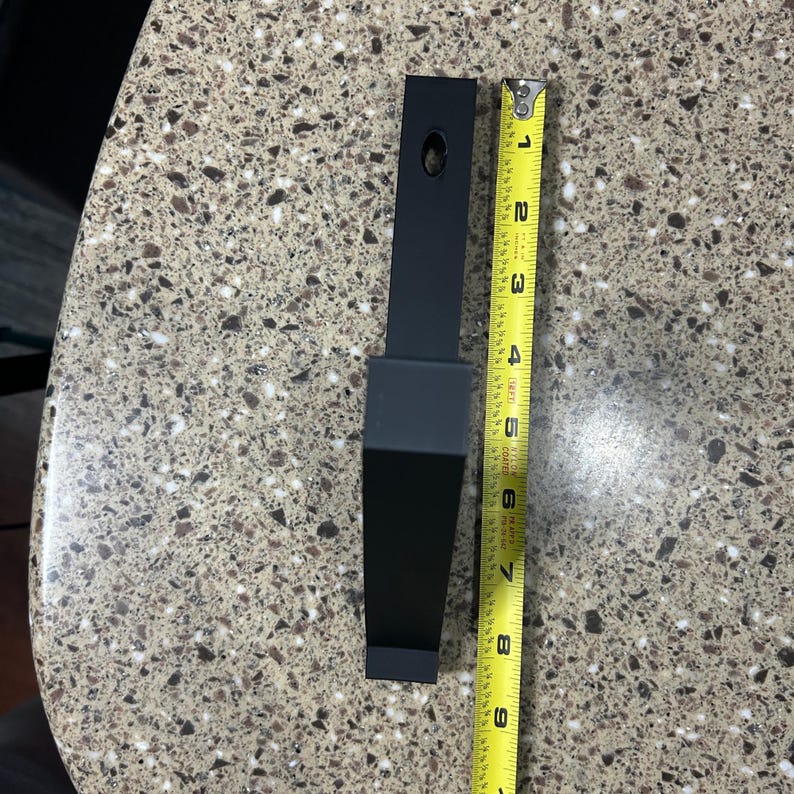 Puede incluir: Un soporte met&aacute;lico negro de forma rectangular, de aproximadamente 20 cm de largo, con un agujero en la parte superior. Una cinta m&eacute;trica amarilla se coloca junto al soporte.