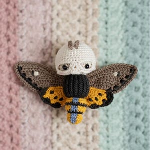 May include: A crocheted moth with a cream-colored head, black eyes, and brown antennae. The moth's wings are brown with black detailing, and its body is black and yellow with blue stripes. The moth is set against a pastel-colored background.