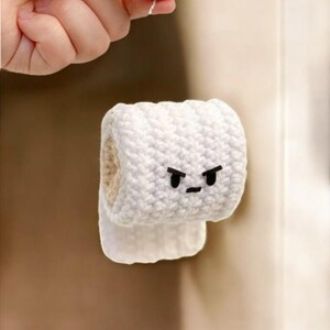 May include: A white, crocheted toilet roll with an angry face, featuring black eyes and a mouth. The handmade item is partially unrolled, showing the inner cardboard tube. The background is blurred, highlighting the craft item.