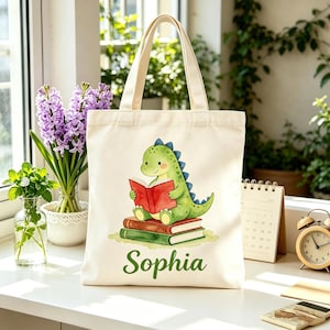 May include: A cream-colored tote bag featuring a green dinosaur reading a red book, sitting atop two other books. The name "Sophia" is printed in green script below the illustration. The bag has cream-colored handles.
