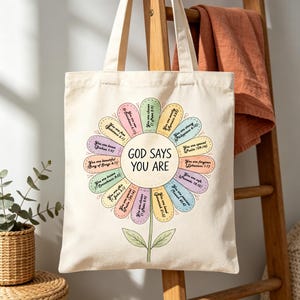 May include: A cream tote bag with a floral design. The flower petals are pastel shades and feature phrases such as "You are loved" and "God says you are." The bag has long handles and a simple design.