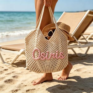 May include: A beige crochet tote bag with the name "Osiris" in pink script. The bag contains a straw hat and sunglasses. The scene is set on a sandy beach with a view of the ocean and a beach chair.