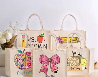 May include: Five beige canvas tote bags with white handles. Each bag features a unique design: a pencil and apple, a floral apple with "Teacher Mrs Smith", a pink bow, a pencil with flowers, and an apple with crayons and "Miss Tracy".