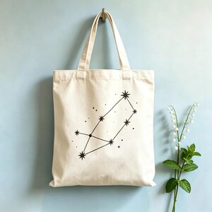 Puede incluir: Una bolsa de tela color crema con un estampado gráfico negro de una constelación. El diseño consta de varias estrellas de ocho puntas conectadas por líneas finas, rodeadas de puntos más pequeños dispersos que representan estrellas.