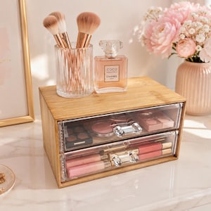 Könnte beinhalten: Ein rechteckiger Make-up-Organizer aus Bambus mit zwei transparenten Acrylschubladen und kristallartigen Knöpfen. Auf der Oberseite befinden sich ein Glasbehälter mit Make-up-Pinseln und ein quadratischer Parfumflakon mit der Aufschrift Coco Chanel.