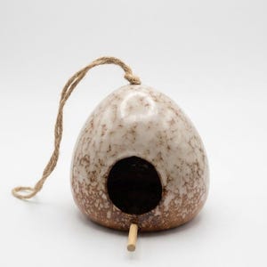 Puede incluir: Una casa para pájaros de cerámica en forma de lágrima con un esmalte moteado en crema y marrón. La estructura presenta un orificio de entrada circular, una pequeña percha de madera que sobresale de la base y un lazo de yute para colgar.