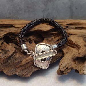 K&ouml;nnte beinhalten: Ein schwarzes, geflochtenes Lederarmband mit einem silbernen Verschluss mit geometrischem Design.
