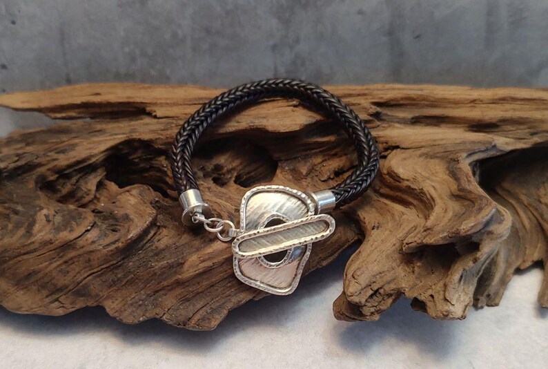K&ouml;nnte beinhalten: Ein schwarzes, geflochtenes Lederarmband mit einem silbernen Verschluss mit geometrischem Design.