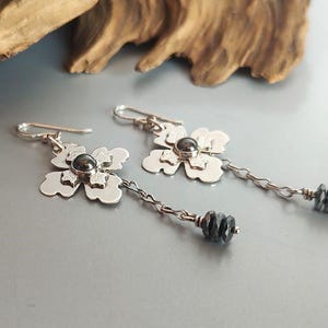Pode incluir: Brincos de prata com flores com um centro de pedra cinza escuro e contas penduradas cinza escuro. Os brincos t&ecirc;m um design floral e est&atilde;o em uma corrente de prata. Fundo cinza claro.