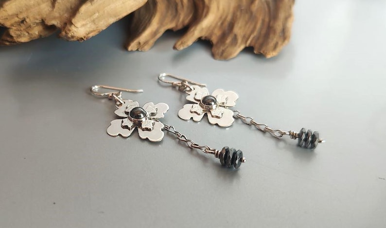 Pode incluir: Brincos de prata com flores com um centro de pedra cinza escuro e contas penduradas cinza escuro. Os brincos t&ecirc;m um design floral e est&atilde;o em uma corrente de prata. Fundo cinza claro.