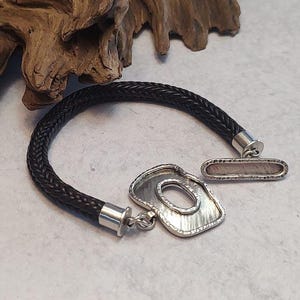 K&ouml;nnte beinhalten: Ein schwarzes geflochtenes Lederarmband mit einem silbernen Verschluss und einem silbernen Anh&auml;nger mit abstraktem Design.