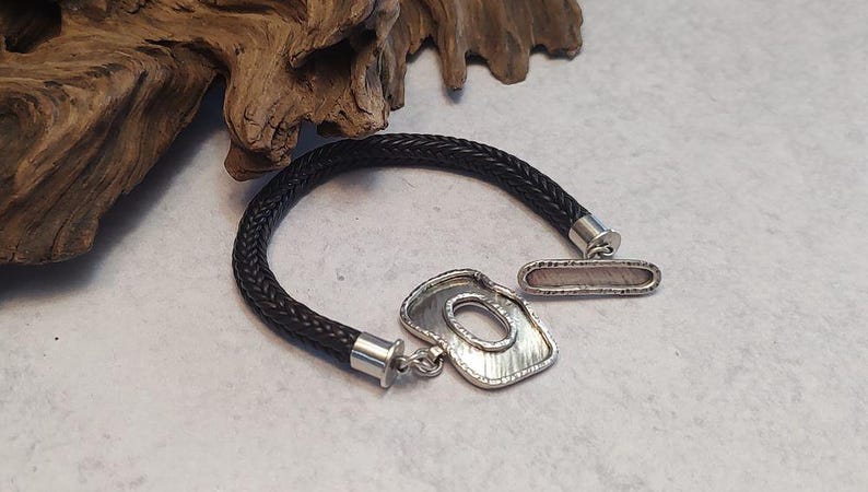 K&ouml;nnte beinhalten: Ein schwarzes geflochtenes Lederarmband mit einem silbernen Verschluss und einem silbernen Anh&auml;nger mit abstraktem Design.