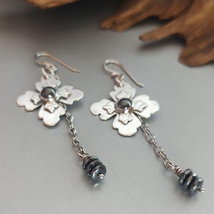 Pode incluir: Brincos de flores prateadas com um centro de pedra cinza escuro e uma corrente de contas cinza escuro. Os brincos t&ecirc;m fecho de gancho e um design floral.