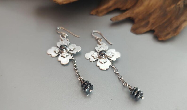 Pode incluir: Brincos de flores prateadas com um centro de pedra cinza escuro e uma corrente de contas cinza escuro. Os brincos t&ecirc;m fecho de gancho e um design floral.