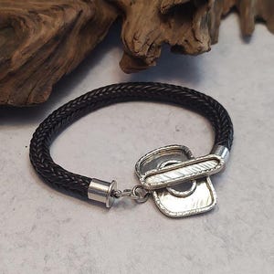 K&ouml;nnte beinhalten: Ein schwarzes geflochtenes Lederarmband mit einem silbernen Verschluss. Der Verschluss ist ein einzigartiges Design mit einer strukturierten Oberfl&auml;che.
