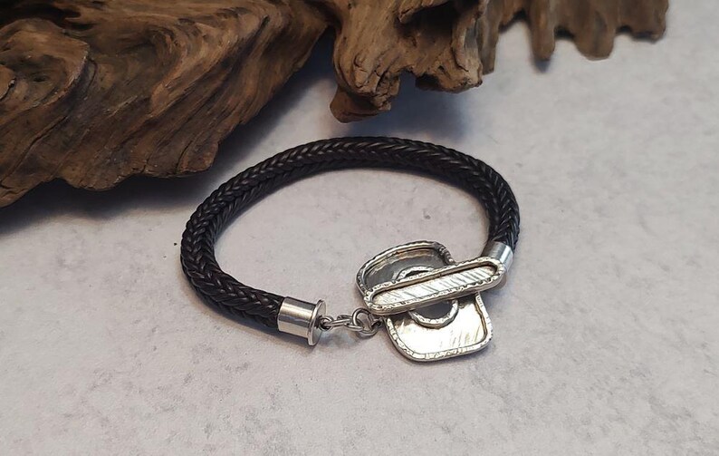 K&ouml;nnte beinhalten: Ein schwarzes geflochtenes Lederarmband mit einem silbernen Verschluss. Der Verschluss ist ein einzigartiges Design mit einer strukturierten Oberfl&auml;che.