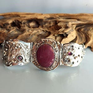 May include: A silver-toned bracelet with a large oval, maroon-colored stone in the center. The bracelet has a textured, ornate design with smaller, round maroon stones. The bracelet is set against a natural wood background.