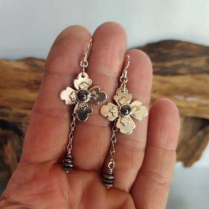 Pode incluir: Brincos de flor prateados com uma pedra escura no centro e detalhes de contas penduradas. Os brincos t&ecirc;m a forma de uma flor de quatro p&eacute;talas. Os brincos est&atilde;o pendurados em um gancho prateado.
